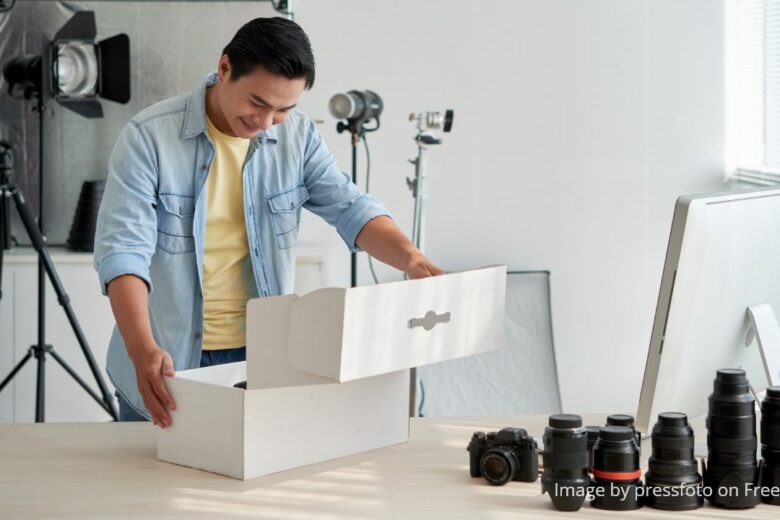 Asian man wearing yellow t-shirt and a light blue collared shirt, smilling while opening up a box of new equipment, probably a camera stabilizer in his photography studio. Image by pressfoto on Feepik.com