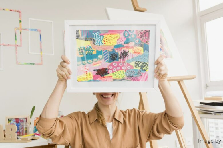 a female artist (in light brown blouse) smiling while holding her colorful piece of artwork above her nose and covering her eyes, in her studio, with an easel stand in the background.