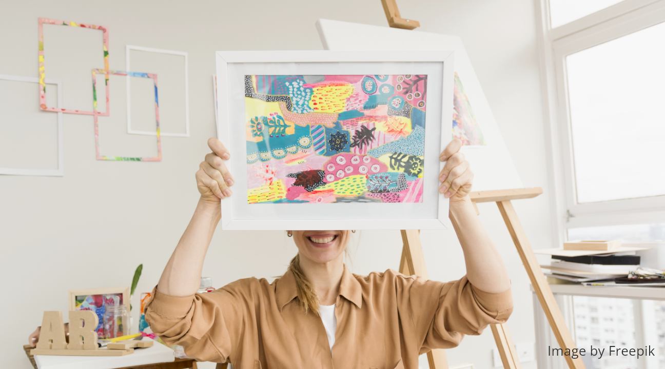 a female artist (in light brown blouse) smiling while holding her colorful piece of artwork above her nose and covering her eyes, in her studio, with an easel stand in the background.