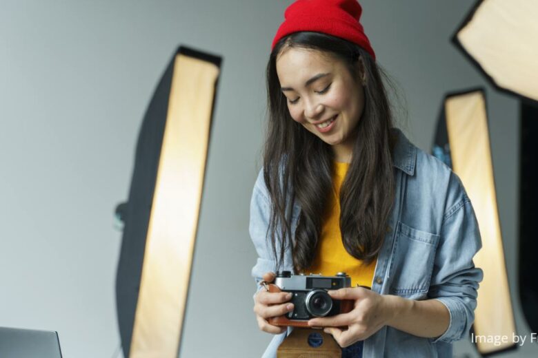 best dslr camera for beginners - Asian young woman with long hair (wearing a red beanie and a denim blouse over a yellow top) smiling while holding a DSLR camera in her photography studio with photography warm white studio lights in the background.