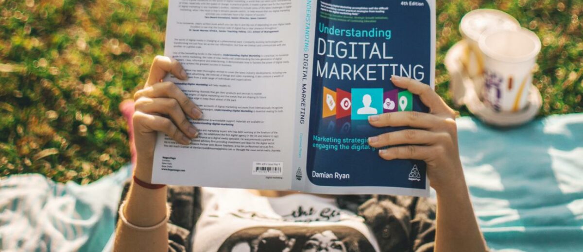 marketing courses - a young lady reading a book about Digital Marketing outdoors with coffee by her side and lying down