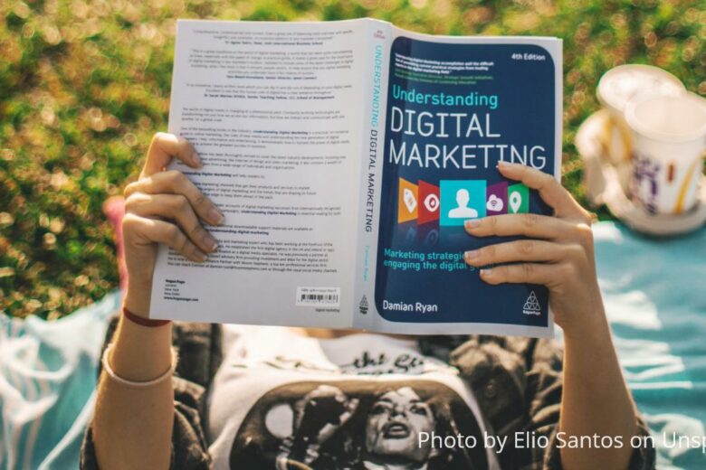 marketing courses - a young lady reading a book about Digital Marketing outdoors with coffee by her side and lying down